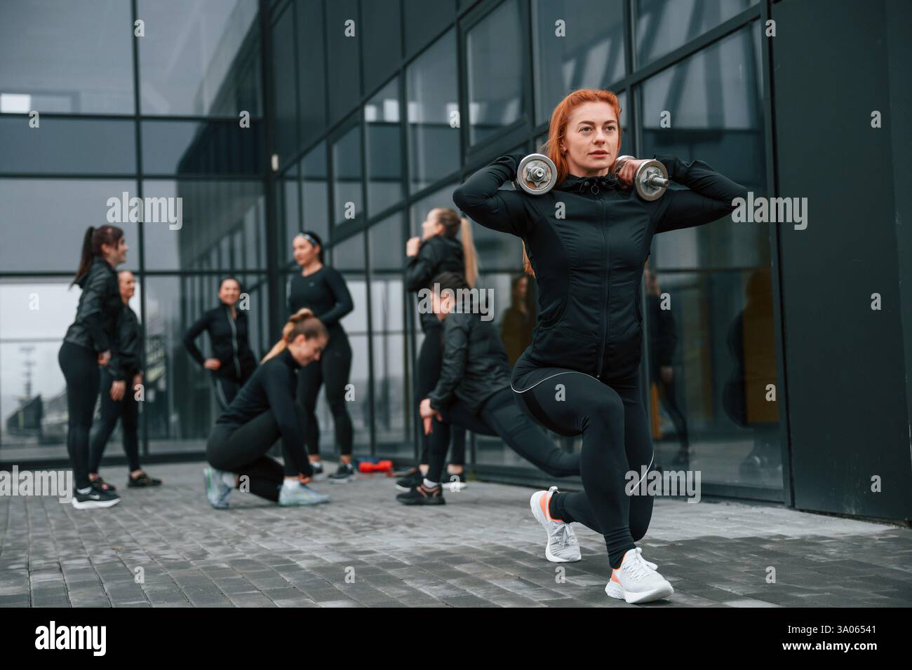La bella donna è in piedi davanti ai suoi amici. Un gruppo di donne sportive si trova all'aperto vicino all'edificio nero. Foto Stock