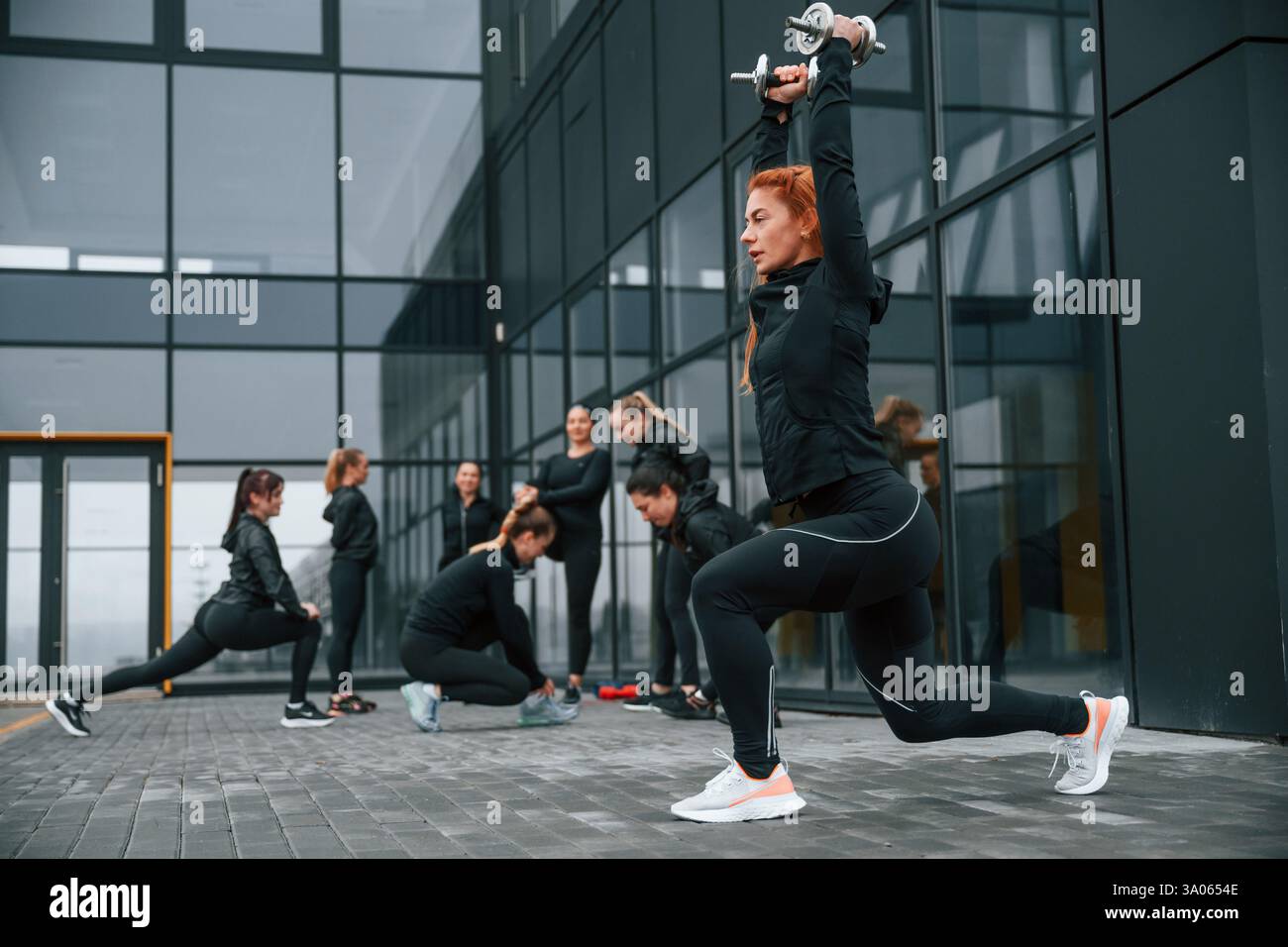 Fare gli squat con i manubri. Un gruppo di donne sportive si trova all'aperto vicino all'edificio nero. Foto Stock