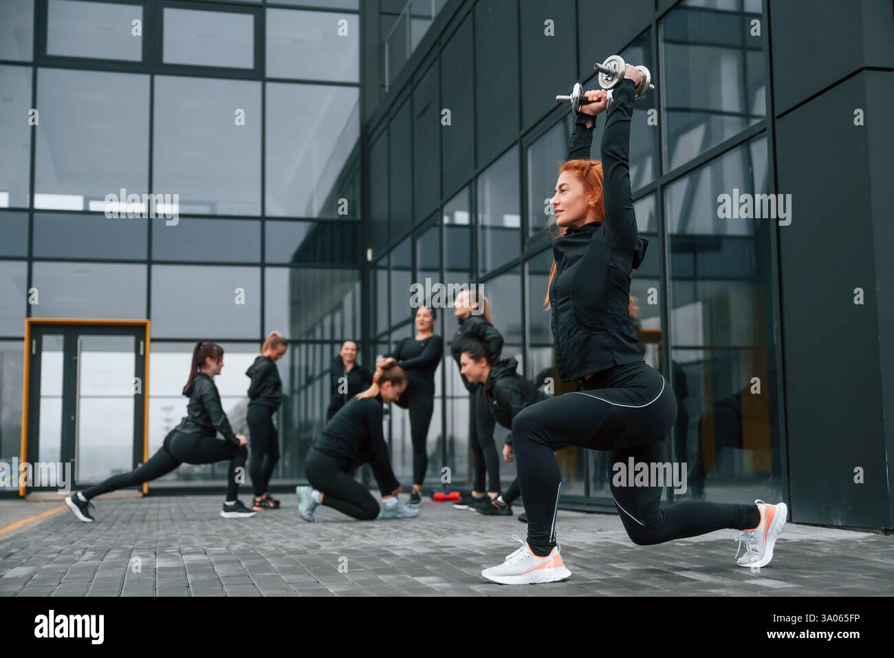 Fare gli squat con i manubri. Un gruppo di donne sportive si trova all'aperto vicino all'edificio nero. Foto Stock