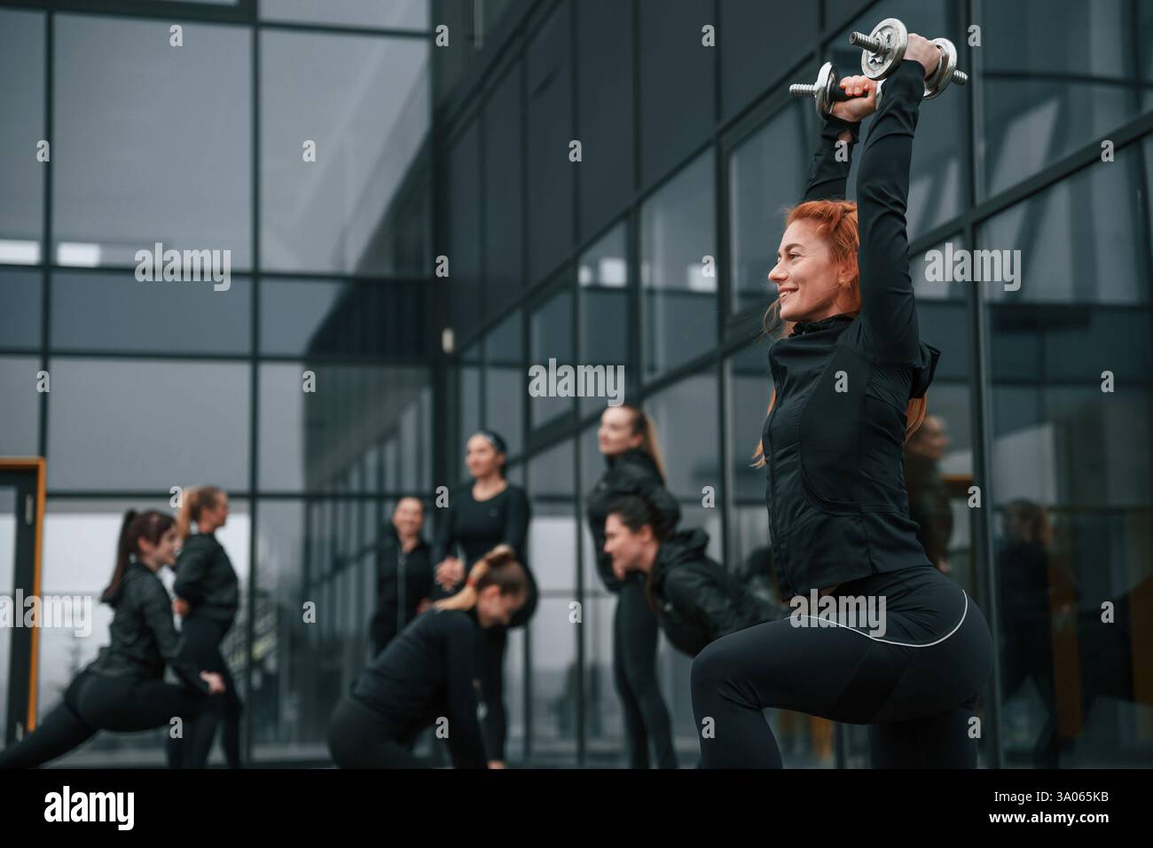 Fare gli squat con i manubri. Un gruppo di donne sportive si trova all'aperto vicino all'edificio nero. Foto Stock