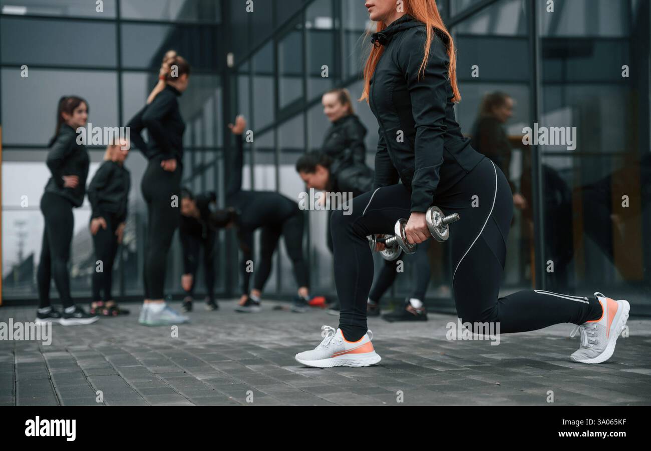 Fare gli squat con i manubri. Un gruppo di donne sportive si trova all'aperto vicino all'edificio nero. Foto Stock