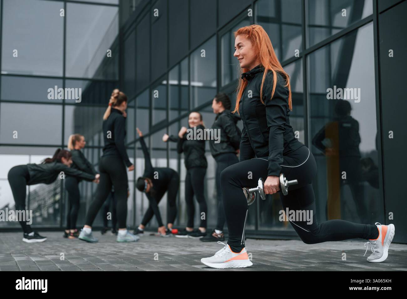 Fare gli squat con i manubri. Un gruppo di donne sportive si trova all'aperto vicino all'edificio nero. Foto Stock