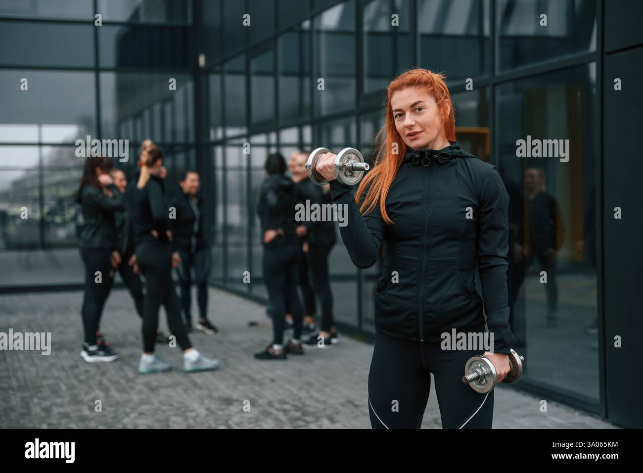 Stare in piedi e tenere i manubri in mano. Un gruppo di donne sportive si trova all'aperto vicino all'edificio nero. Foto Stock