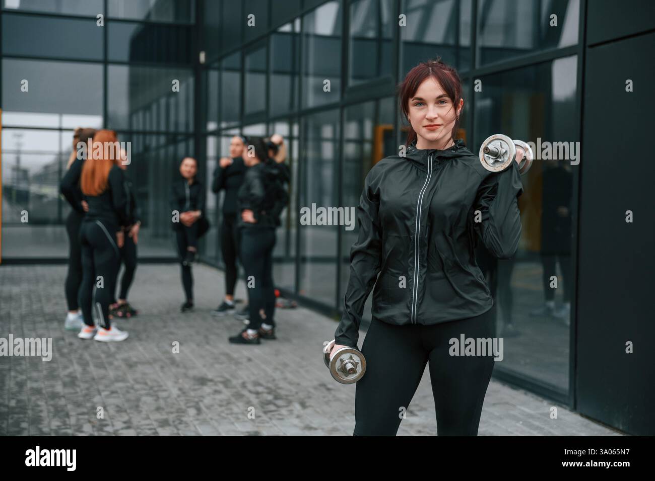 Stare in piedi e tenere i manubri in mano. Un gruppo di donne sportive si trova all'aperto vicino all'edificio nero. Foto Stock