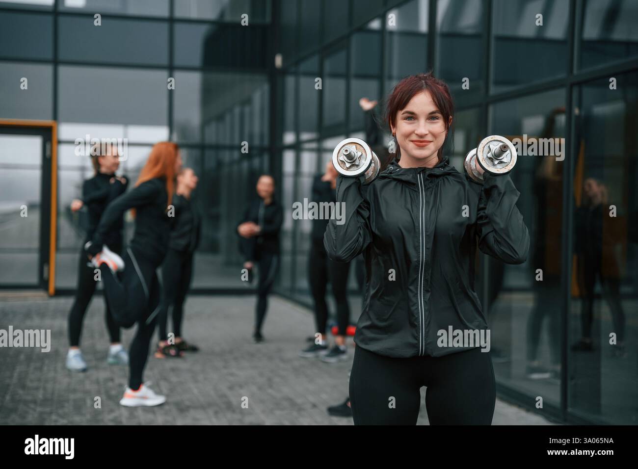 Stare in piedi e tenere i manubri in mano. Un gruppo di donne sportive si trova all'aperto vicino all'edificio nero. Foto Stock