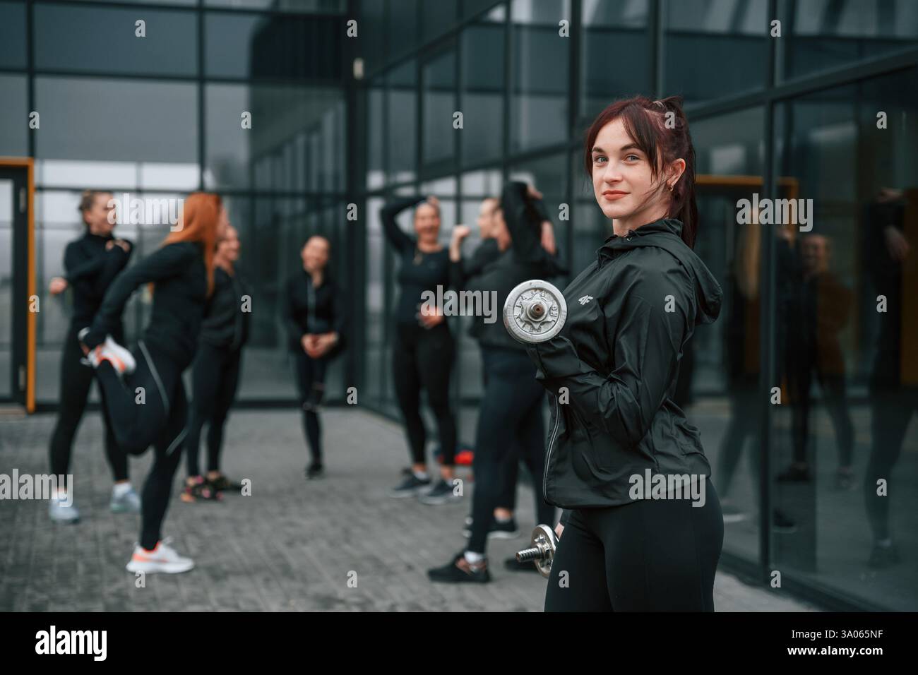 Stare in piedi e tenere i manubri in mano. Un gruppo di donne sportive si trova all'aperto vicino all'edificio nero. Foto Stock