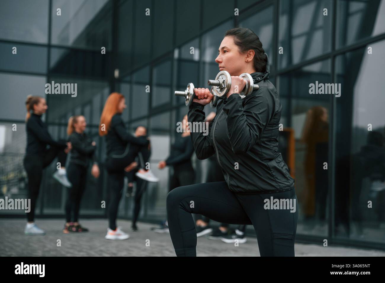 Vista laterale. Fare gli squat e tenere i manubri. Un gruppo di donne sportive si trova all'aperto vicino all'edificio nero. Foto Stock