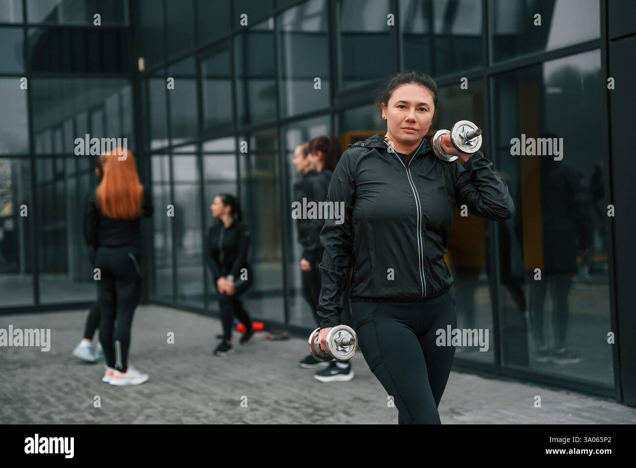 Stare in piedi e tenere i manubri in mano. Un gruppo di donne sportive si trova all'aperto vicino all'edificio nero. Foto Stock