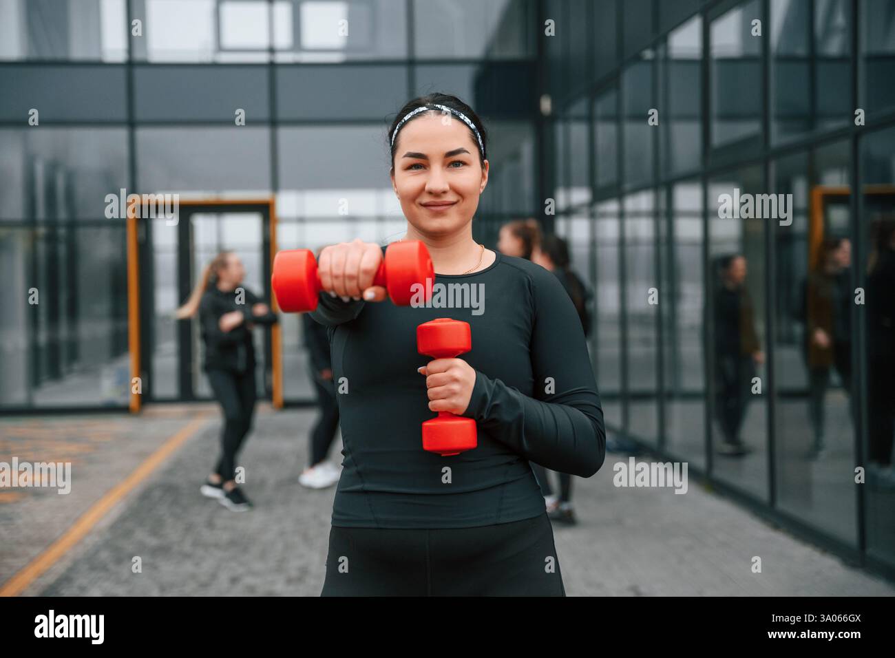 Sorridere, fare esercizi usando i manubri. Un gruppo di donne sportive si trova all'aperto vicino all'edificio nero. Foto Stock