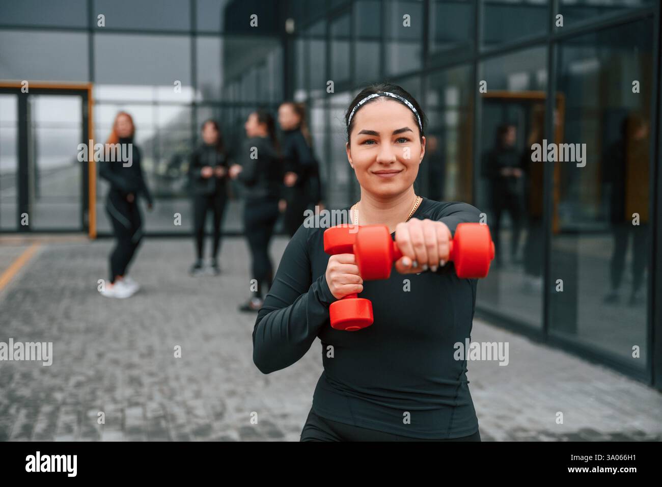 Sorridere, fare esercizi usando i manubri. Un gruppo di donne sportive si trova all'aperto vicino all'edificio nero. Foto Stock