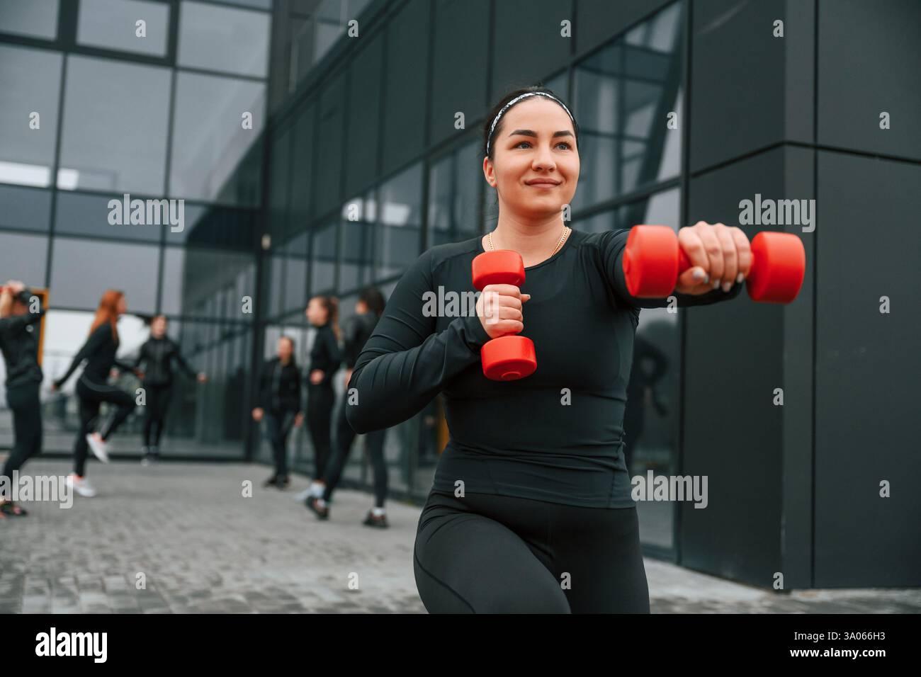 Sorridere, fare esercizi usando i manubri. Un gruppo di donne sportive si trova all'aperto vicino all'edificio nero. Foto Stock