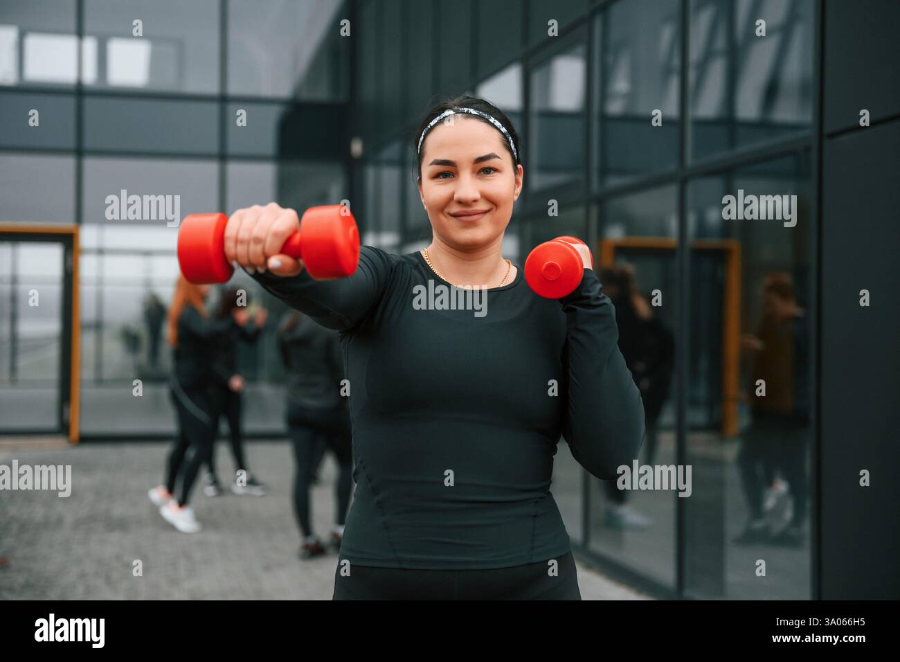 Sorridere, fare esercizi usando i manubri. Un gruppo di donne sportive si trova all'aperto vicino all'edificio nero. Foto Stock