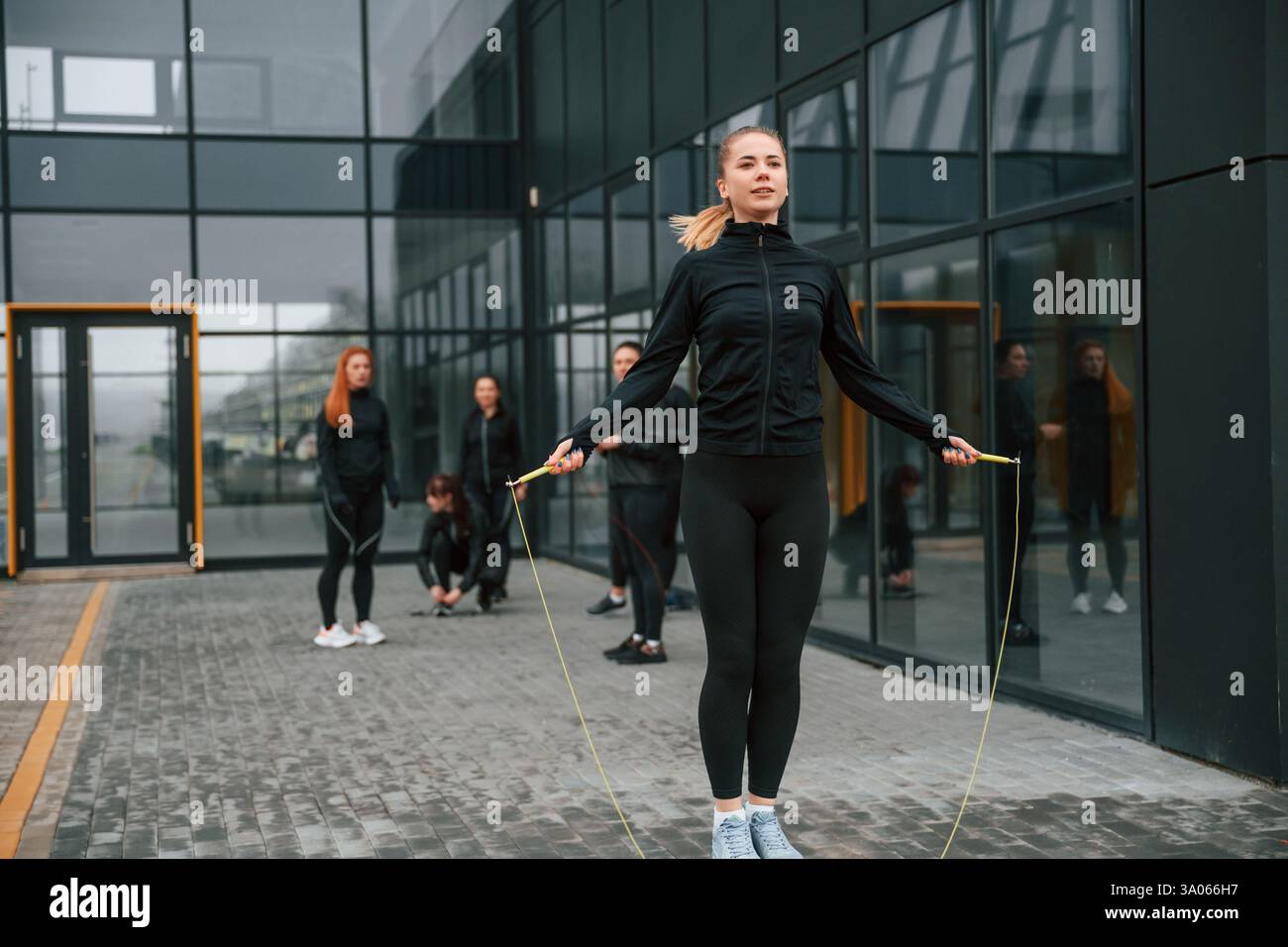 Fare esercizi usando la corda per saltare. Un gruppo di donne sportive si trova all'aperto vicino all'edificio nero. Foto Stock
