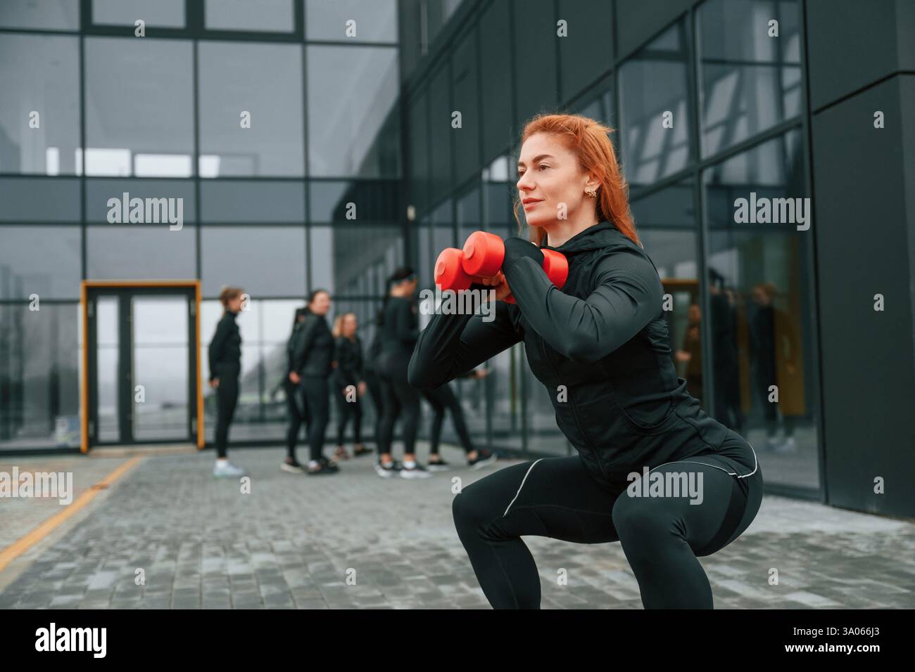 Fare esercizi con i manubri. Un gruppo di donne sportive si trova all'aperto vicino all'edificio nero. Foto Stock