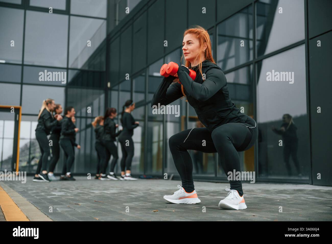 Fare esercizi con i manubri. Un gruppo di donne sportive si trova all'aperto vicino all'edificio nero. Foto Stock