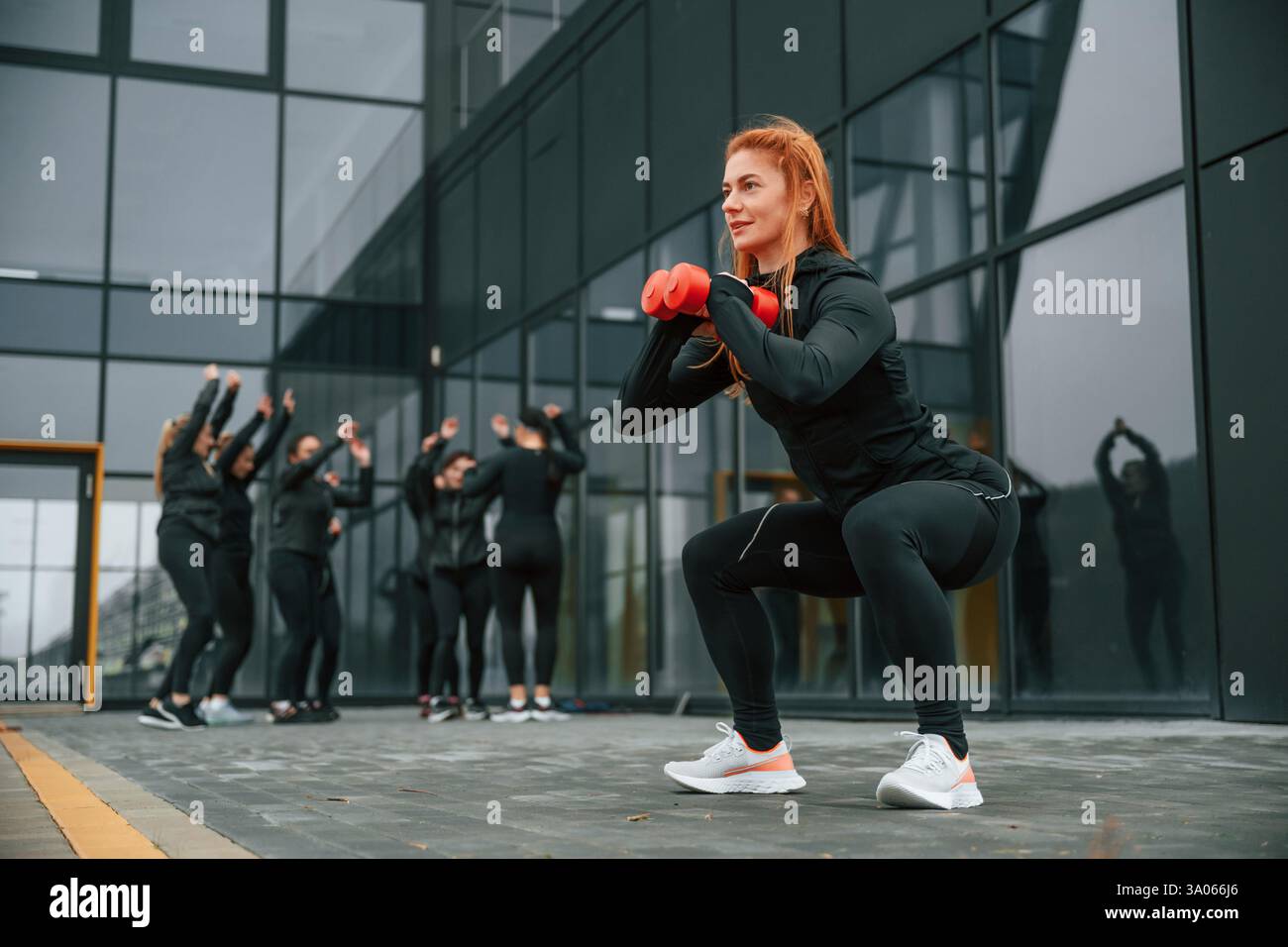 Fare esercizi con i manubri. Un gruppo di donne sportive si trova all'aperto vicino all'edificio nero. Foto Stock