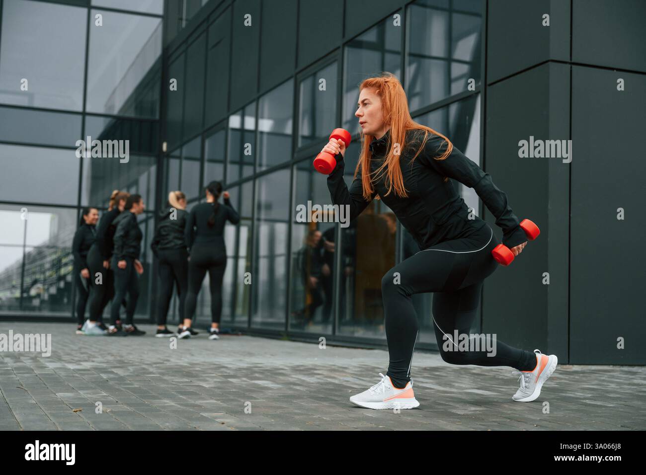 Fare esercizi con i manubri. Un gruppo di donne sportive si trova all'aperto vicino all'edificio nero. Foto Stock