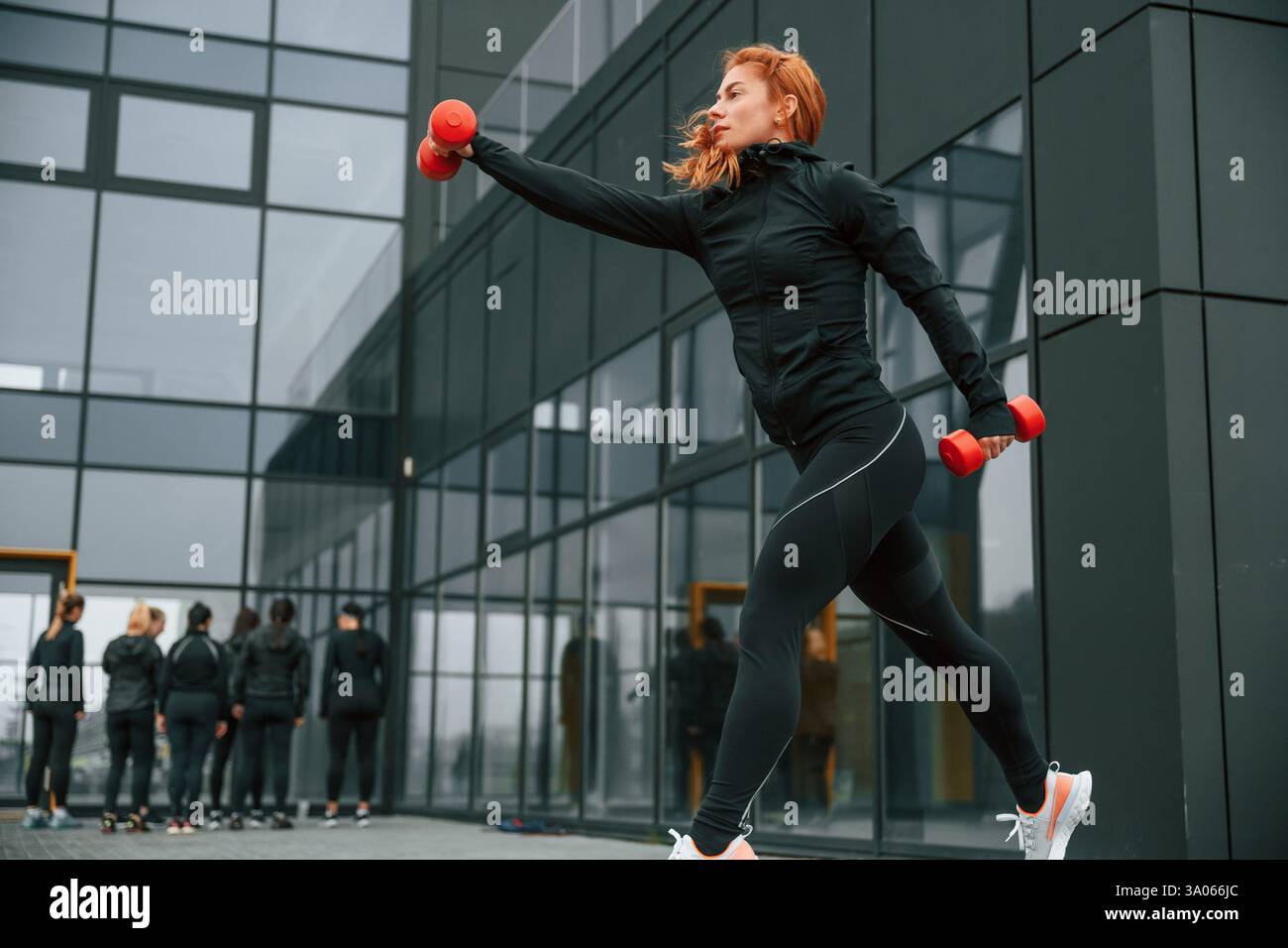 Fare esercizi con i manubri. Un gruppo di donne sportive si trova all'aperto vicino all'edificio nero. Foto Stock