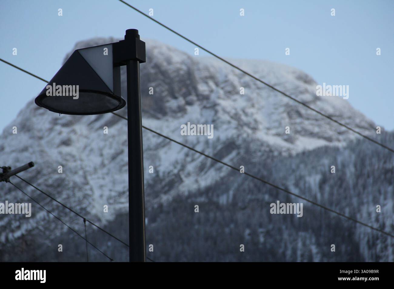 Moderno lampione nero silouettato contro un imponente picco montuoso coperto di neve con linee elettriche sopraelevate in un paesaggio invernale freddo. Foto Stock