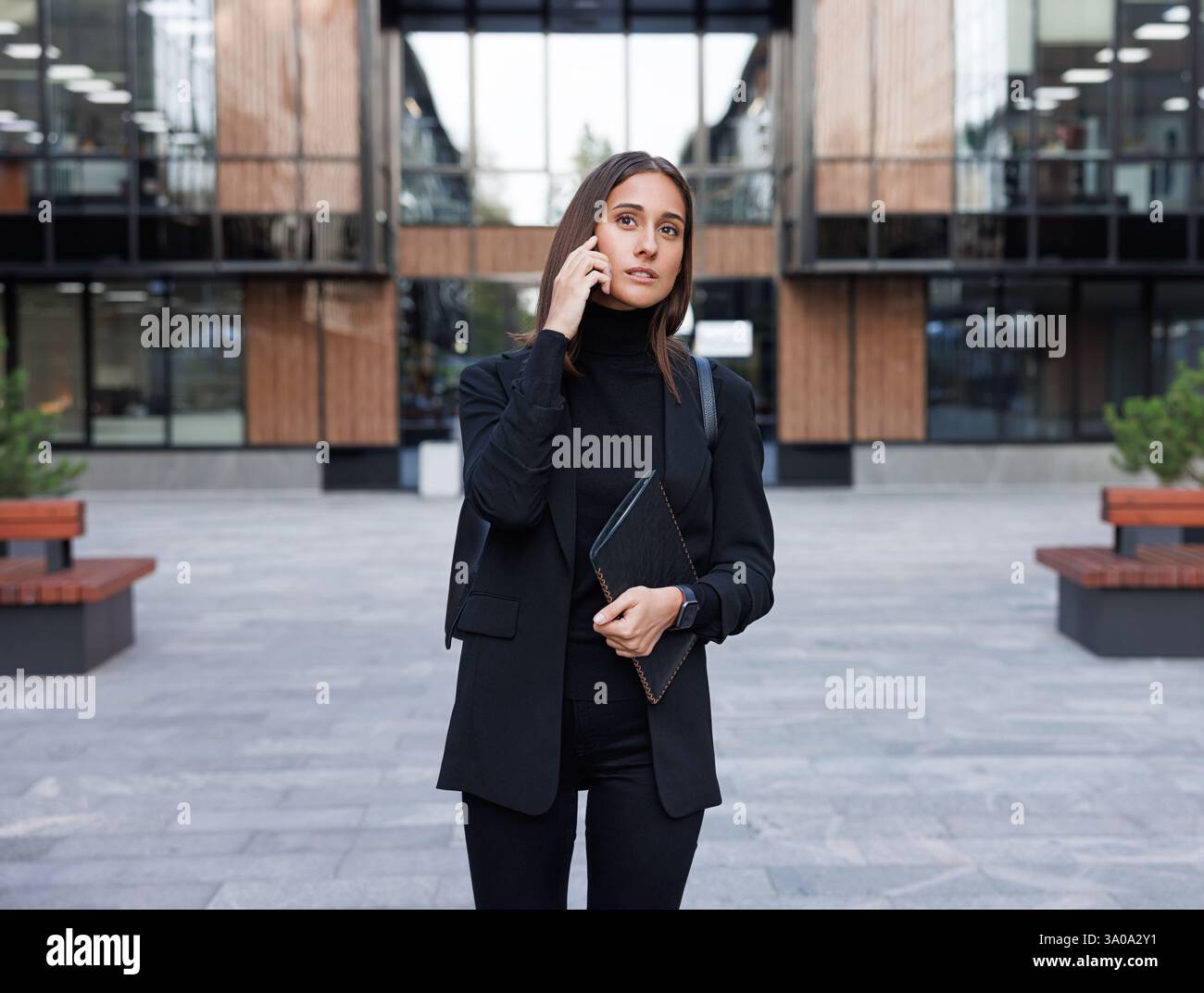 Giovane imprenditrice che indossa abiti eleganti e formali neri con colletto alto che effettua una telefonata nel cortile di campagna di un edificio per uffici Foto Stock