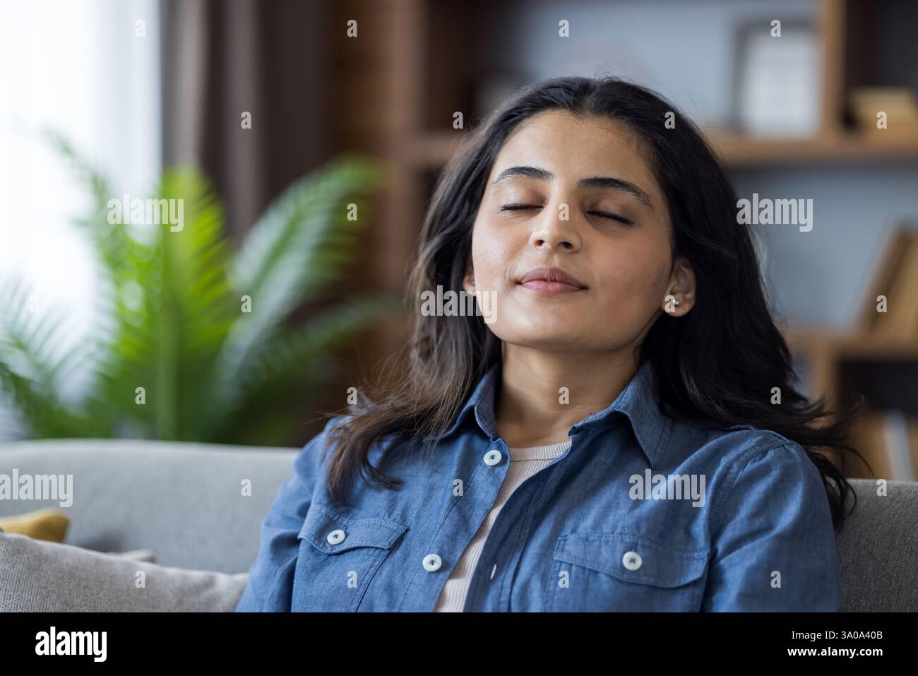 Giovane donna che si rilassa a casa sul divano, gli occhi chiusi, pratica meditazione e consapevolezza per alleviare lo stress in un accogliente soggiorno. Foto Stock