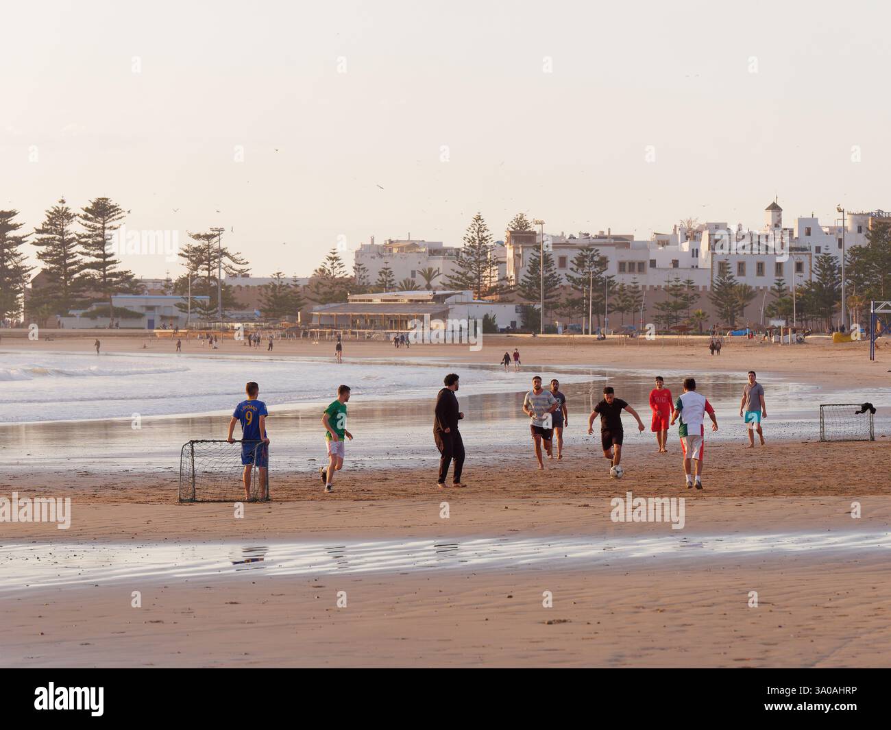 Gli adolescenti giocano a calcio su una spiaggia di sabbia bagnata con la storica Medina alle spalle di Essaouira. 2 marzo 2025 Foto Stock