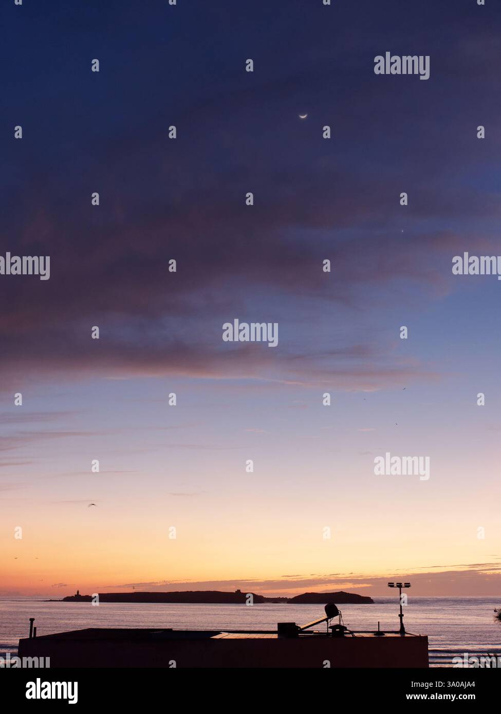 Luna nel cielo scuro al tramonto con il cielo rosso e l'isola sottostante sul mare Atlantico con tetto in primo piano a Essaouira. 2 marzo 2025 Foto Stock