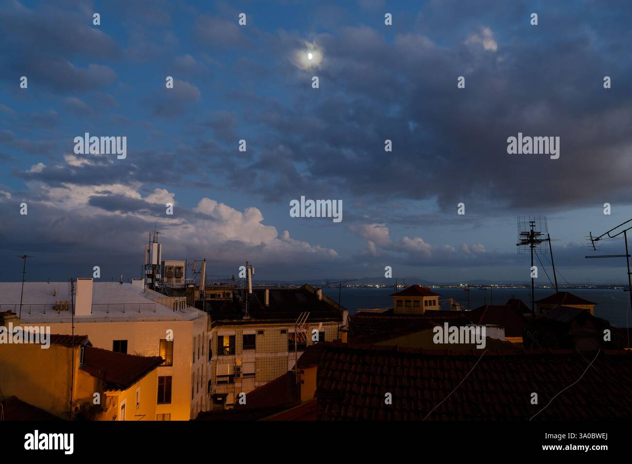 Una serata illuminata dalla luna sui tetti di Alfama, dove le luci della città e le spettacolari nuvole fanno da cornice al tranquillo fiume Tago Foto Stock