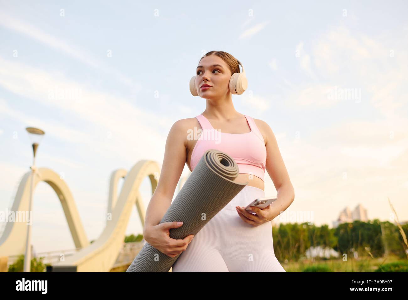 Una giovane donna sicura di sé tiene il suo tappetino da yoga, pronto per una sessione all'aperto. Foto Stock