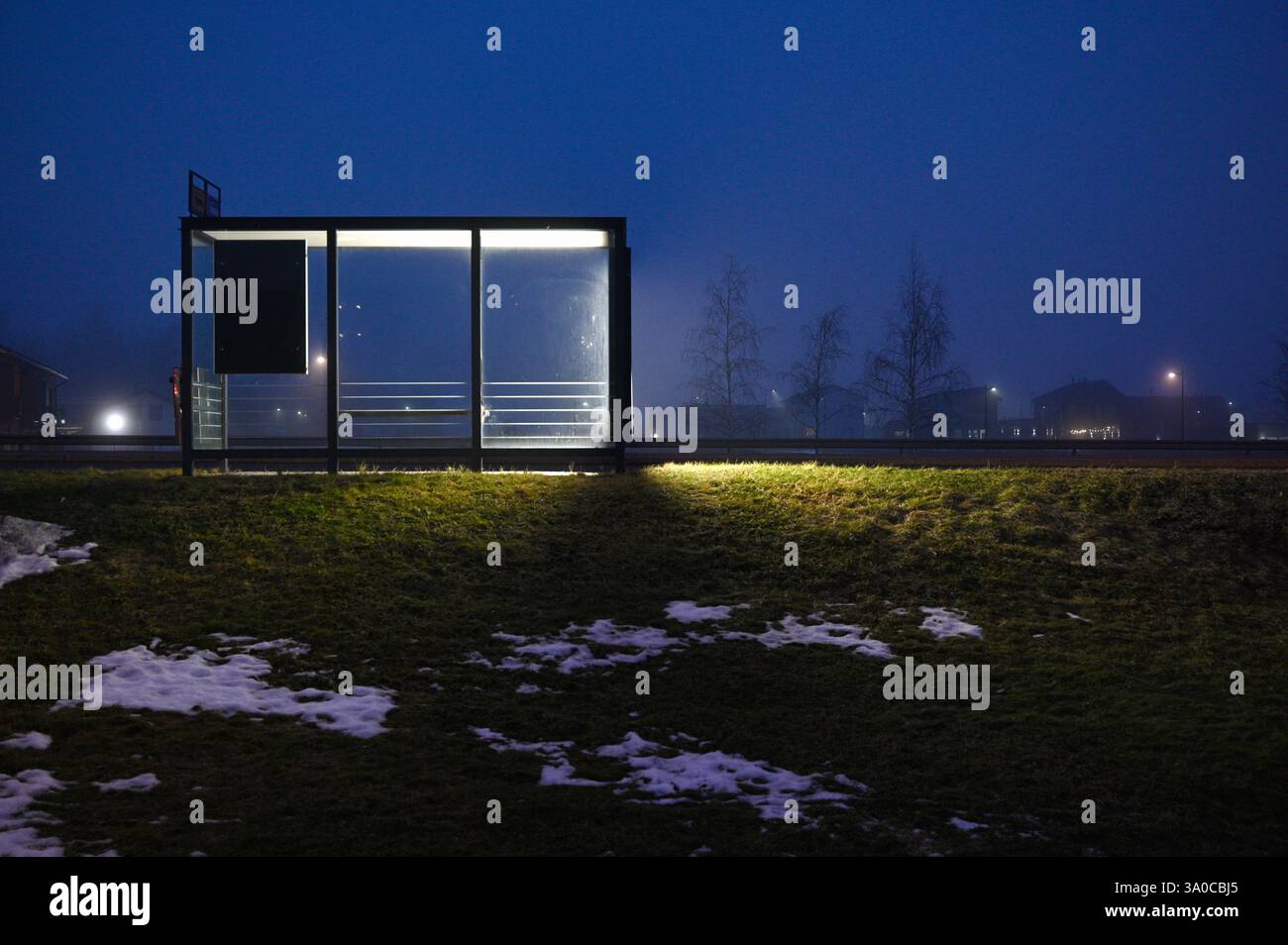 Una fermata minimalista dell'autobus illuminata da pannelli luminosi in una serata nebbiosa, immersa in un tranquillo paesaggio suburbano. Macchie di neve a terra e luce soffusa Foto Stock