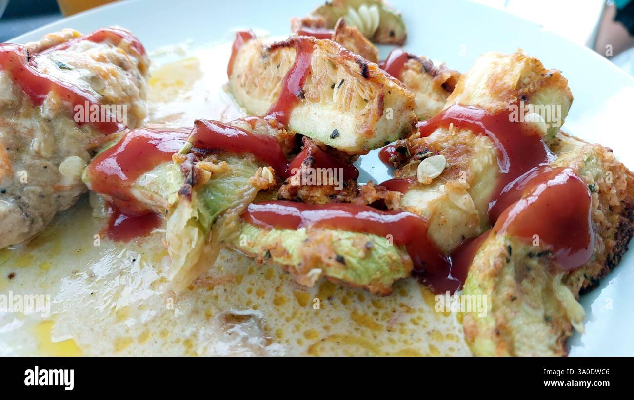Le lancette di zucchine grigliate riposano invitantemente su un piatto bianco pulito, ricoperto da vivace ketchup rosso, che mostra una deliziosa creazione culinaria perfetta per Foto Stock