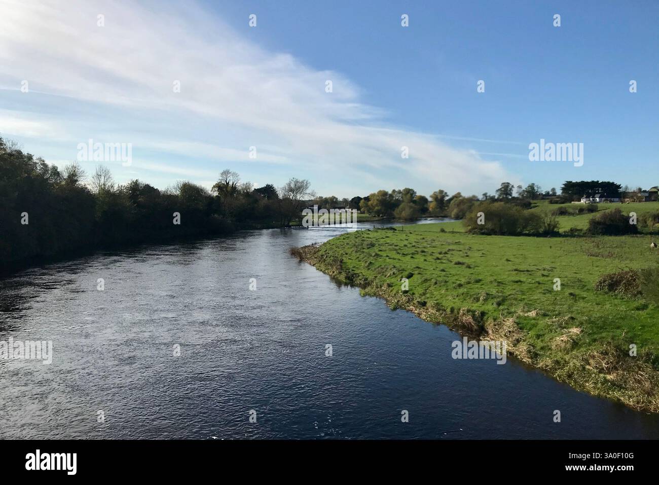 Una tranquilla scena della campagna irlandese caratterizzata da un fiume che scorre dolcemente, lussureggianti campi verdi e un cielo blu limpido. Foto Stock
