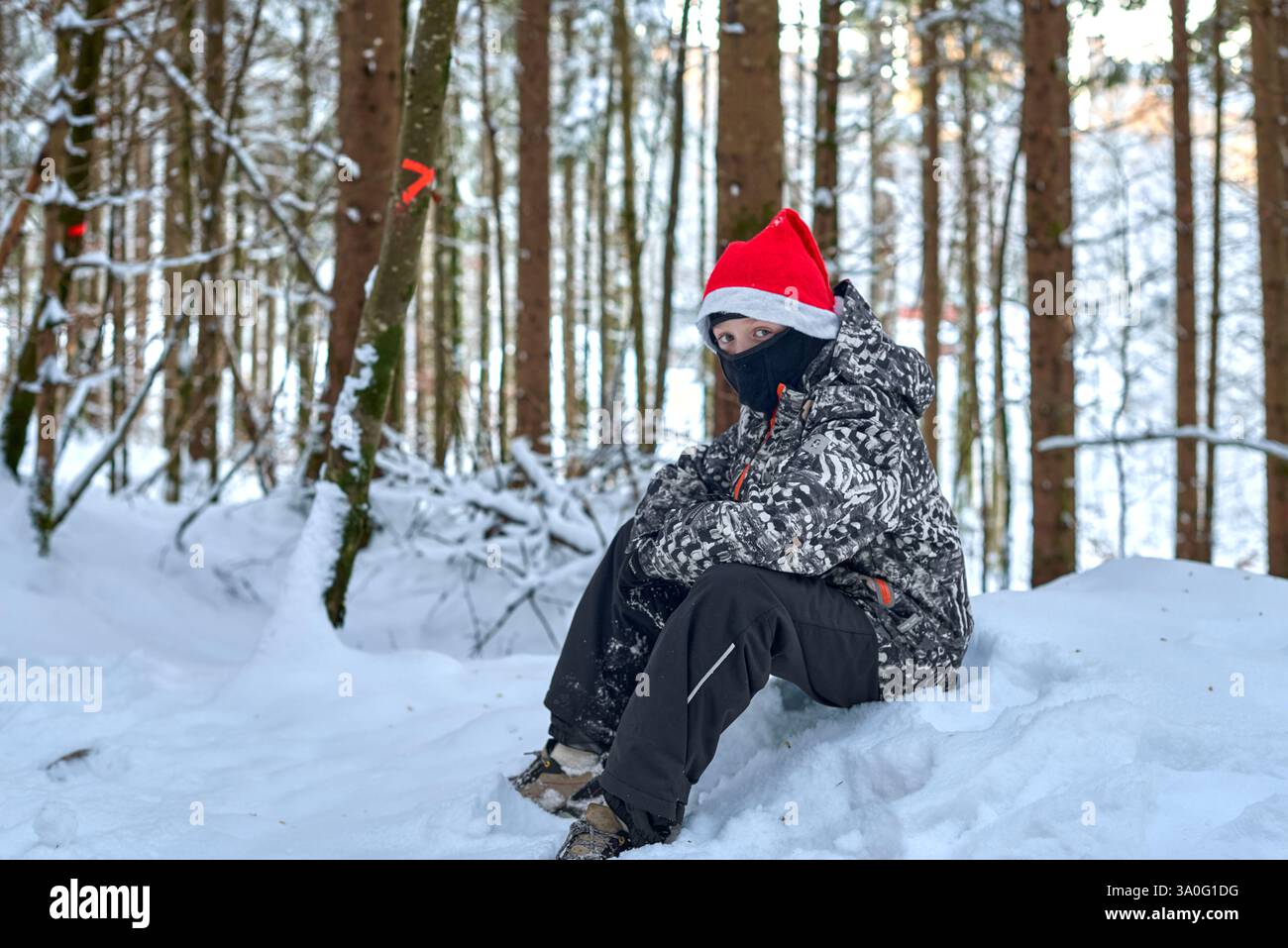 Suggestivo ritratto invernale che raffigura un bambino vestito con abiti invernali mimetici e un cappello di Babbo Natale rosso festoso, in piedi con cura su una copertura neve Foto Stock