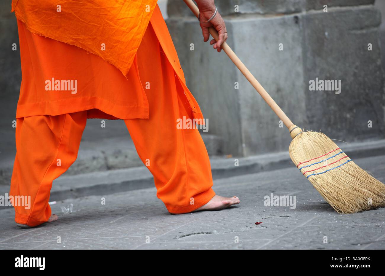 Monaco indiano che indossa abiti arancioni che spazza la strada con una scopa durante una processione, mostrando dedizione alla pulizia e alla spiritualità all'interno del Foto Stock