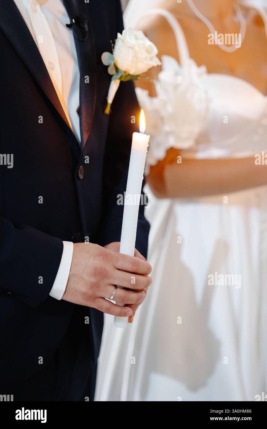 Primo piano delle mani dello sposo tenendo una candela accesa durante una cerimonia nuziale, simboleggiando l'unità e la speranza Foto Stock