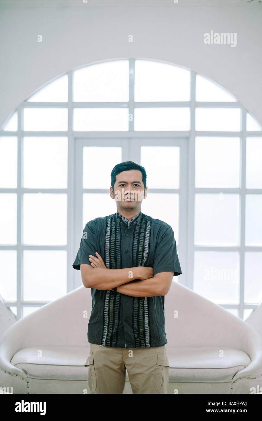 Un uomo indonesiano fiducioso si erge con le braccia incrociate in una camera luminosa e moderna. L'uomo indossa una camicia casual e pantaloni cargo. Ha un neutrale Foto Stock
