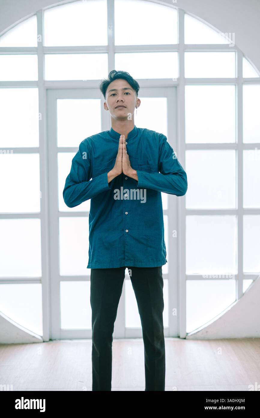 Un giovane contemplativo in casa, vestito con un abbigliamento formale informale, si unisce alle mani in una postura pacifica, esprimendo gratitudine e consapevolezza Foto Stock