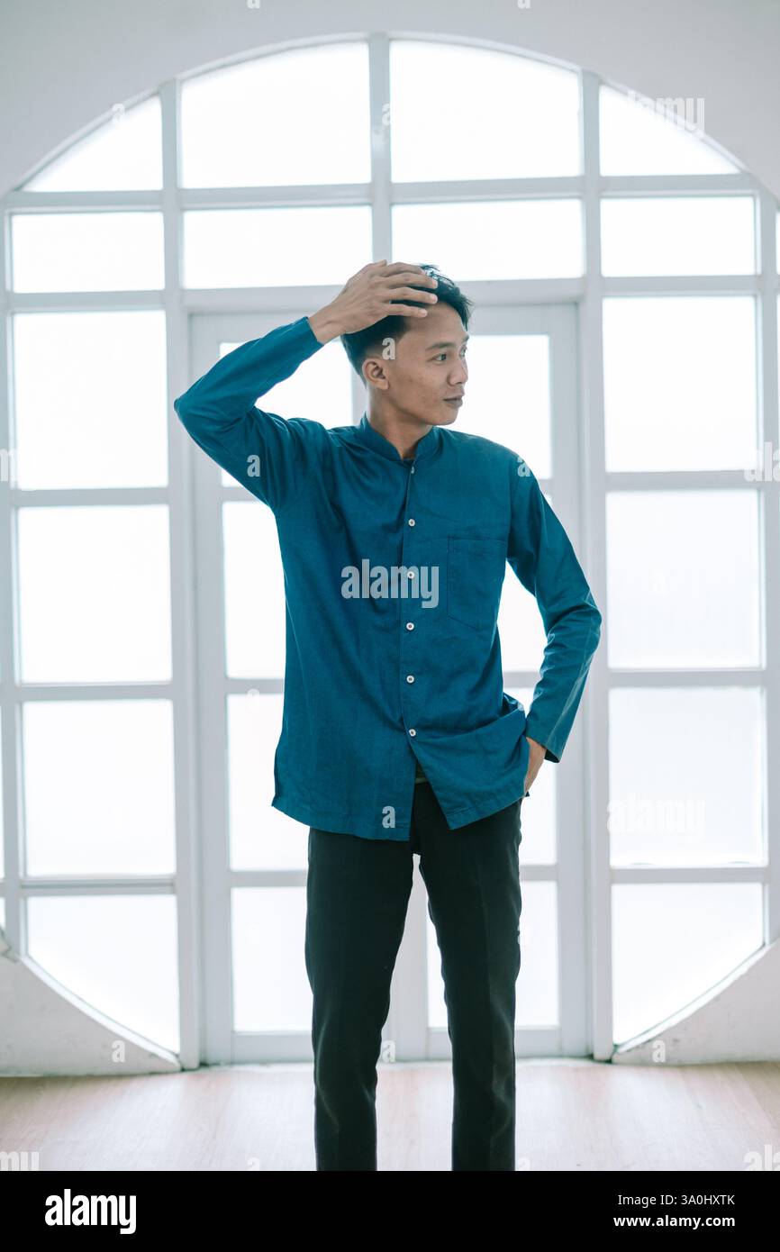 Un giovane uomo con un elegante abbigliamento casual si fa notare in una posa premurosa. Scattata contro una finestra luminosa, l'immagine trasmette un senso di eleganza moderna. Sta in piedi Foto Stock