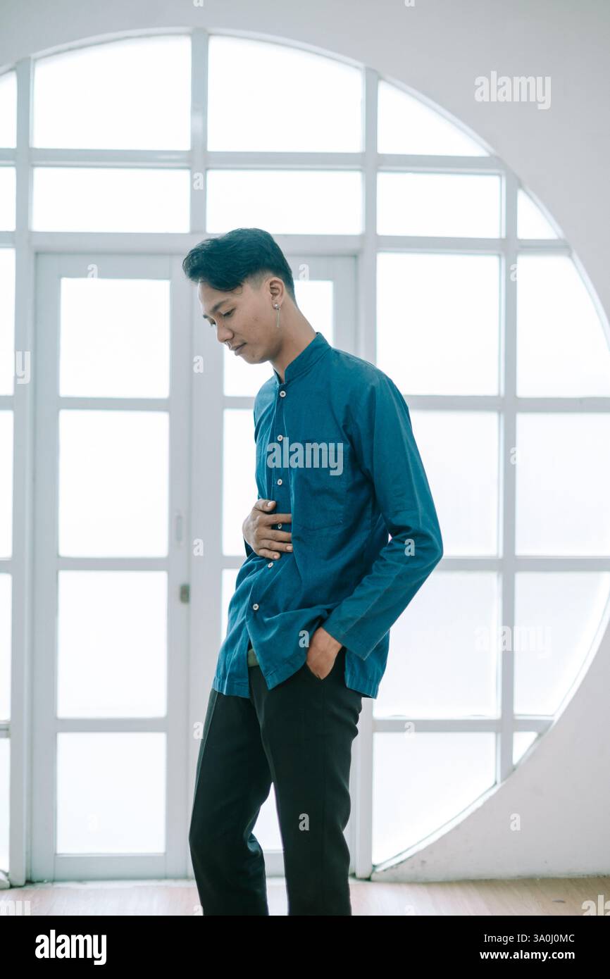 Un giovane uomo si staglia con cura in interni, indossando una camicia casual blu e pantaloni neri. Trasuda uno stile moderno, la sua posa contemplativa cattura Foto Stock