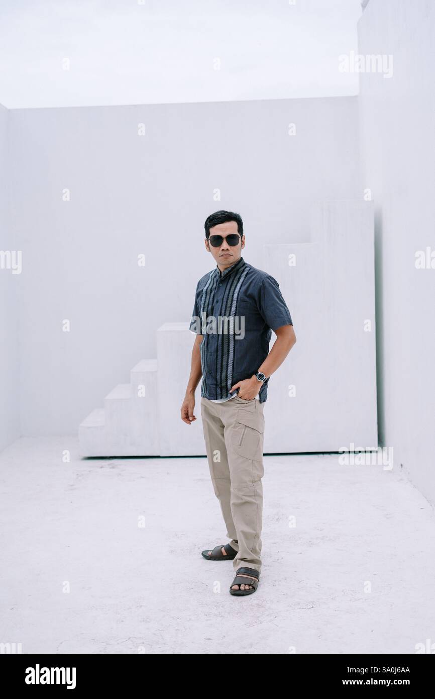 Un uomo moderno posa con sicurezza all'interno di uno spazio geometrico bianco nitido. Il suo outfit elegante e casual, completo di occhiali da sole e orologio, parla con il suo refi Foto Stock