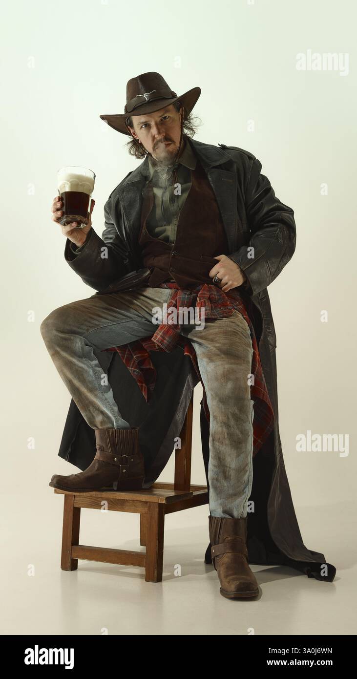 Uomo brutale vestito da cowboy seduto sulla sedia, fumando e tenendo un bicchiere di birra in schiuma scura. Foto Stock