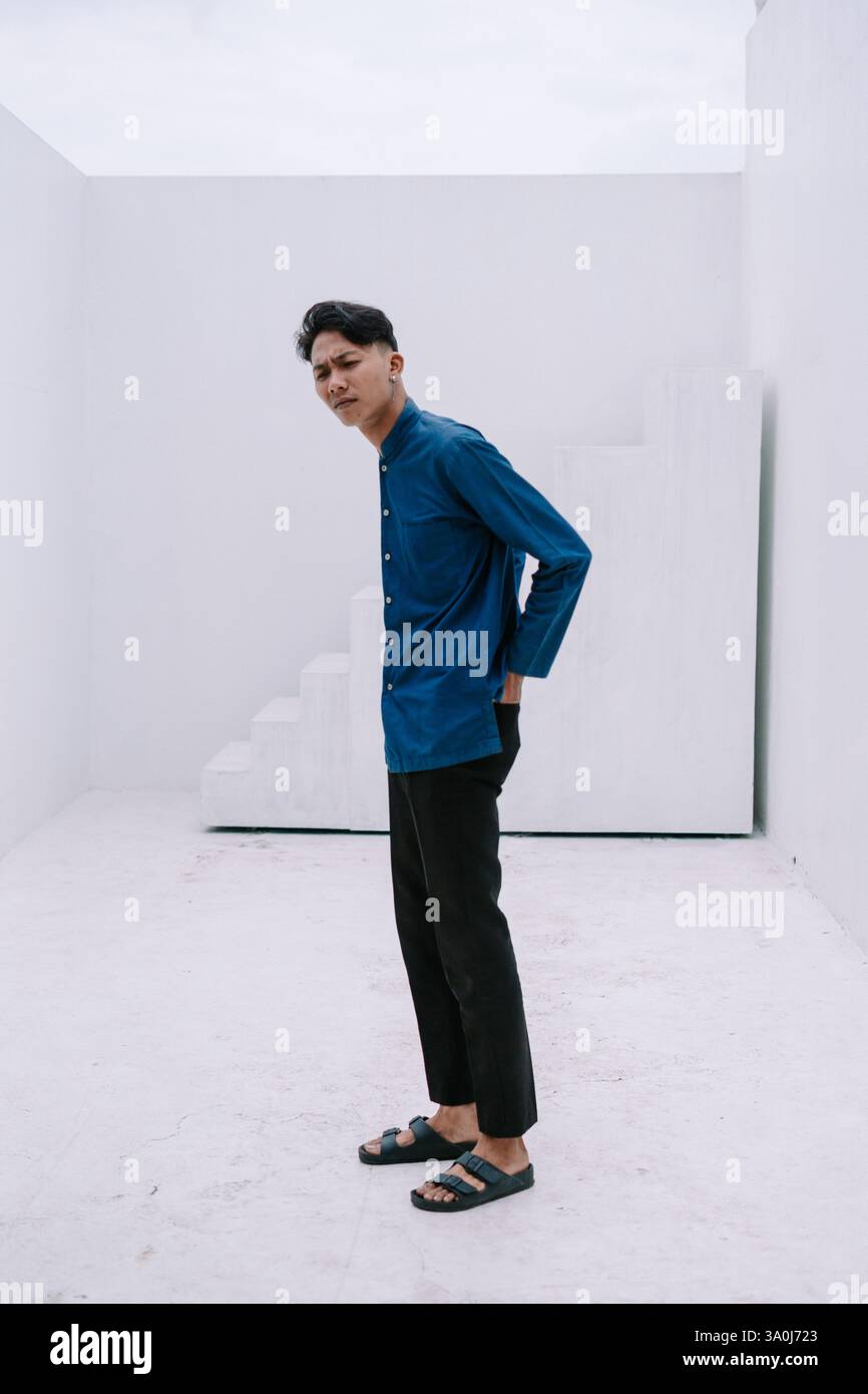 Un giovane uomo alla moda si posa in un ambiente minimalista in bianco studio, trasudando fiducia in un ambiente informale. È vestito con una camicia blu e pantaloni neri, reflecti Foto Stock