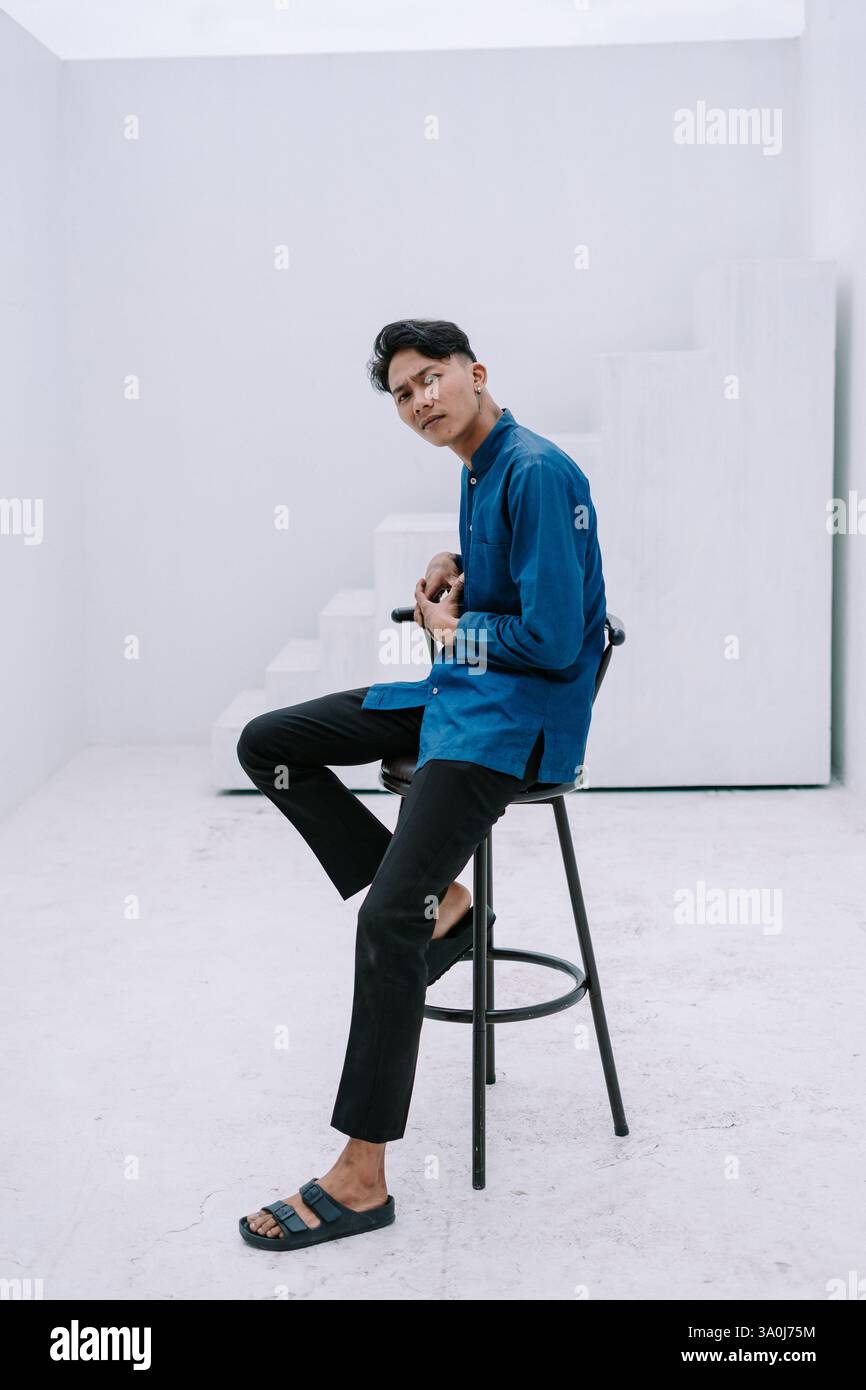Un giovane asiatico siede casualmente su uno sgabello minimalista. È vestito con un abbigliamento alla moda e si mette in posa con cura, guardando con fiducia nel Foto Stock