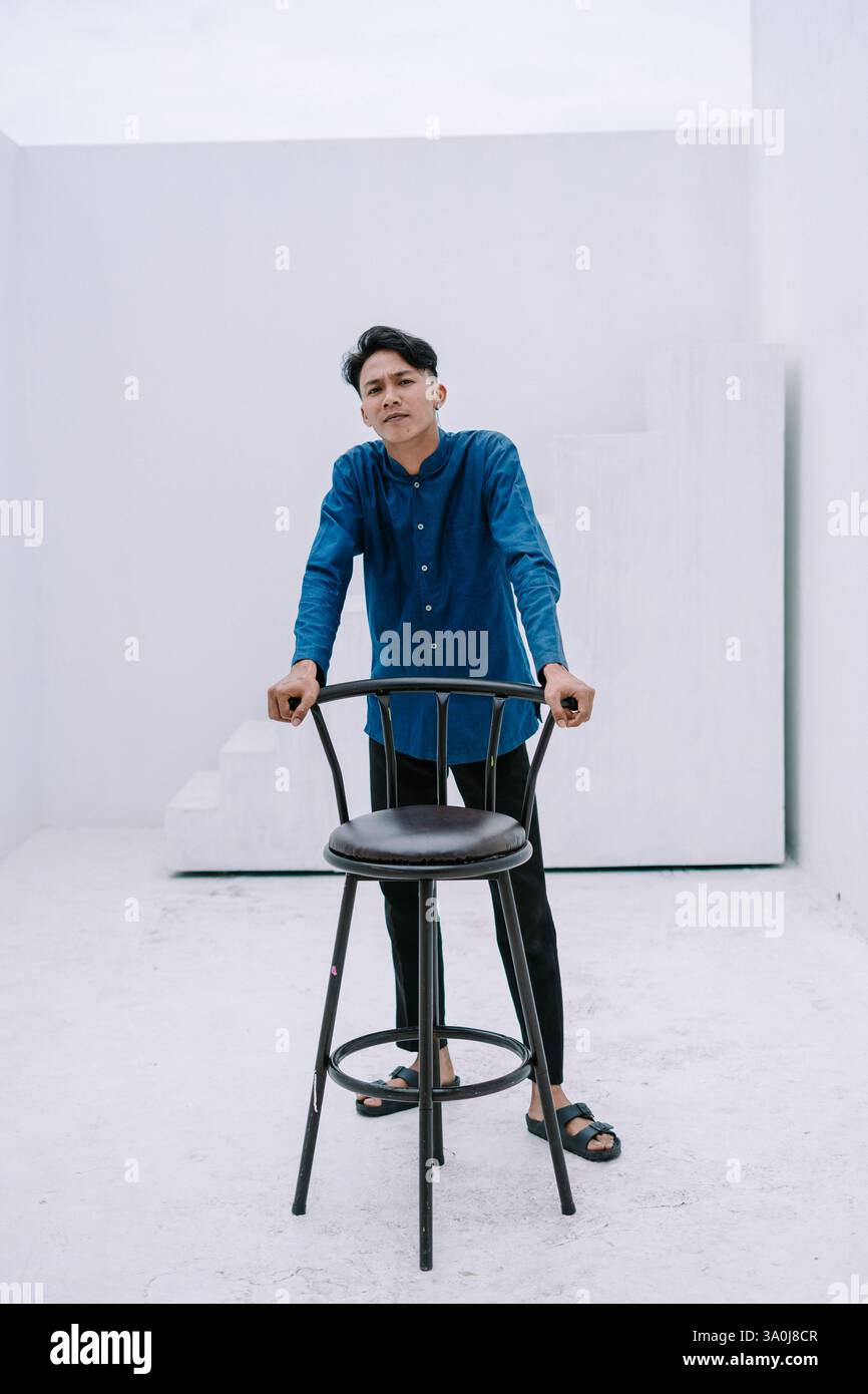 Ritratto di un giovane attento in un abbigliamento casual appoggiato su una sedia all'interno di uno studio minimalista. Sfondo pulito e luminoso con forme geometriche. Suita Foto Stock