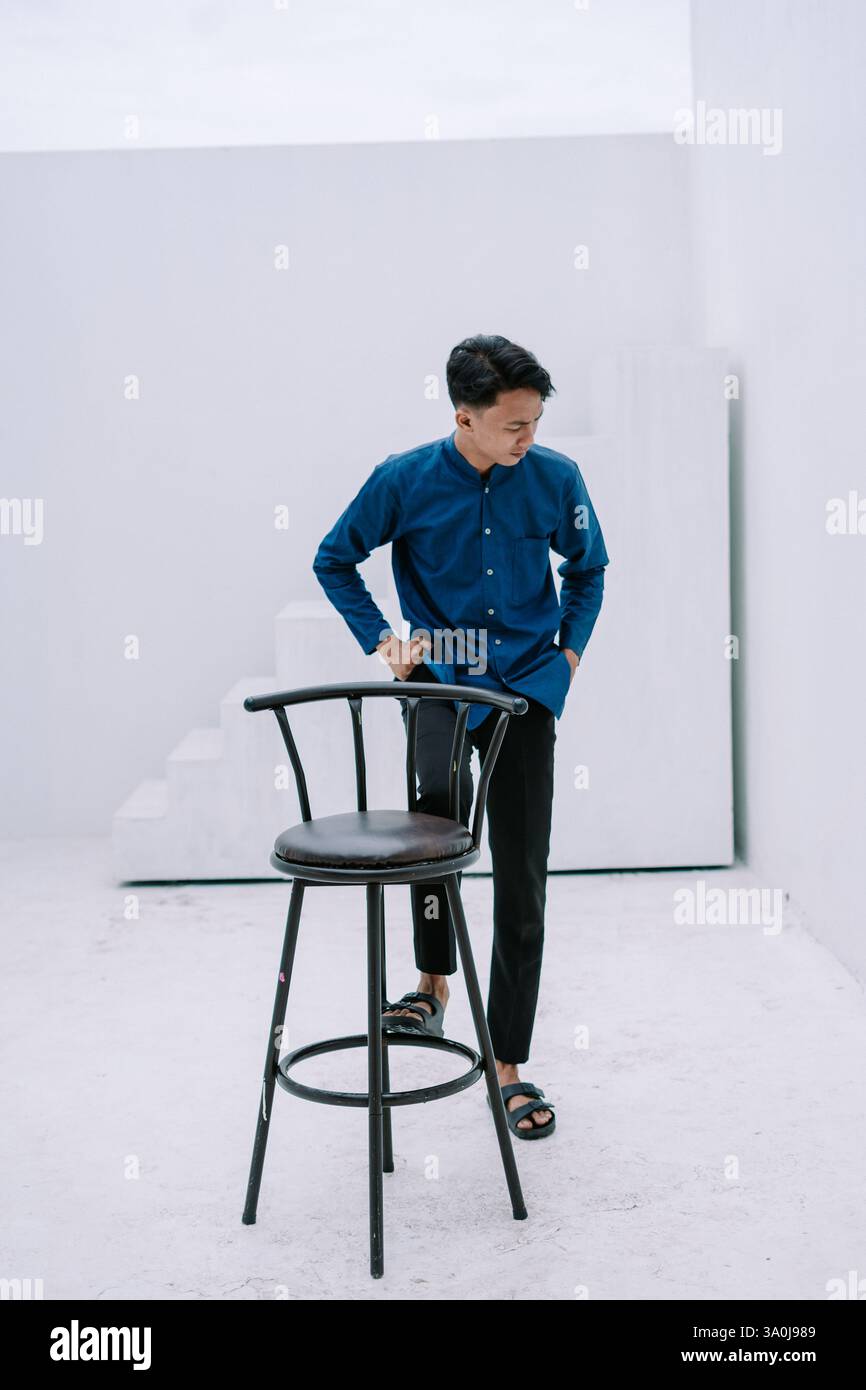 Un giovane uomo che indossa un'elegante camicia blu e pantaloni neri si appoggia a uno sgabello da bar nero in uno studio bianco minimalista. Trasuda un moderno e. Foto Stock