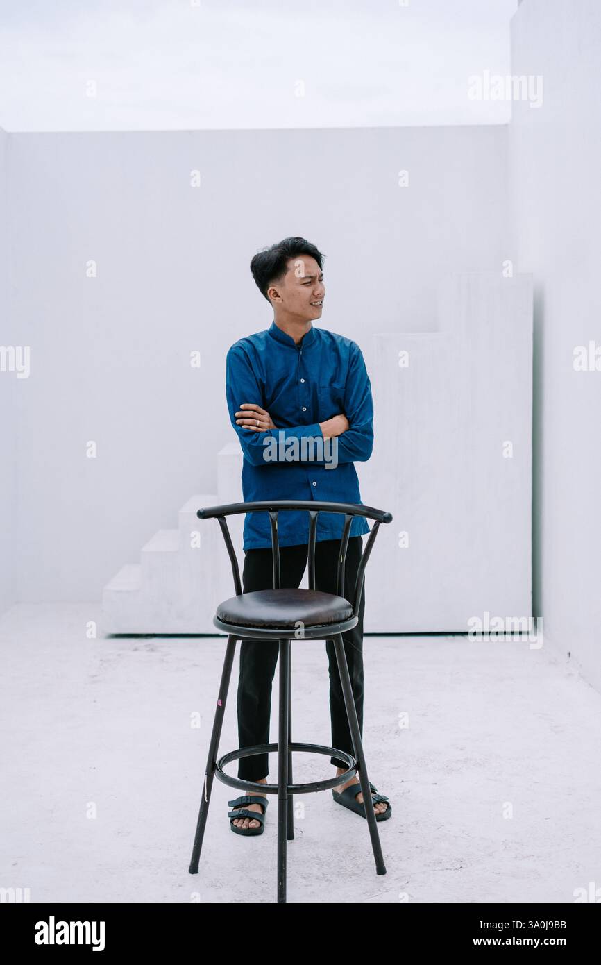 Un giovane asiatico che indossa una camicia blu in piedi con sicurezza con le braccia incrociate in uno studio bianco minimalista. Si sta inclinando casualmente contro Foto Stock