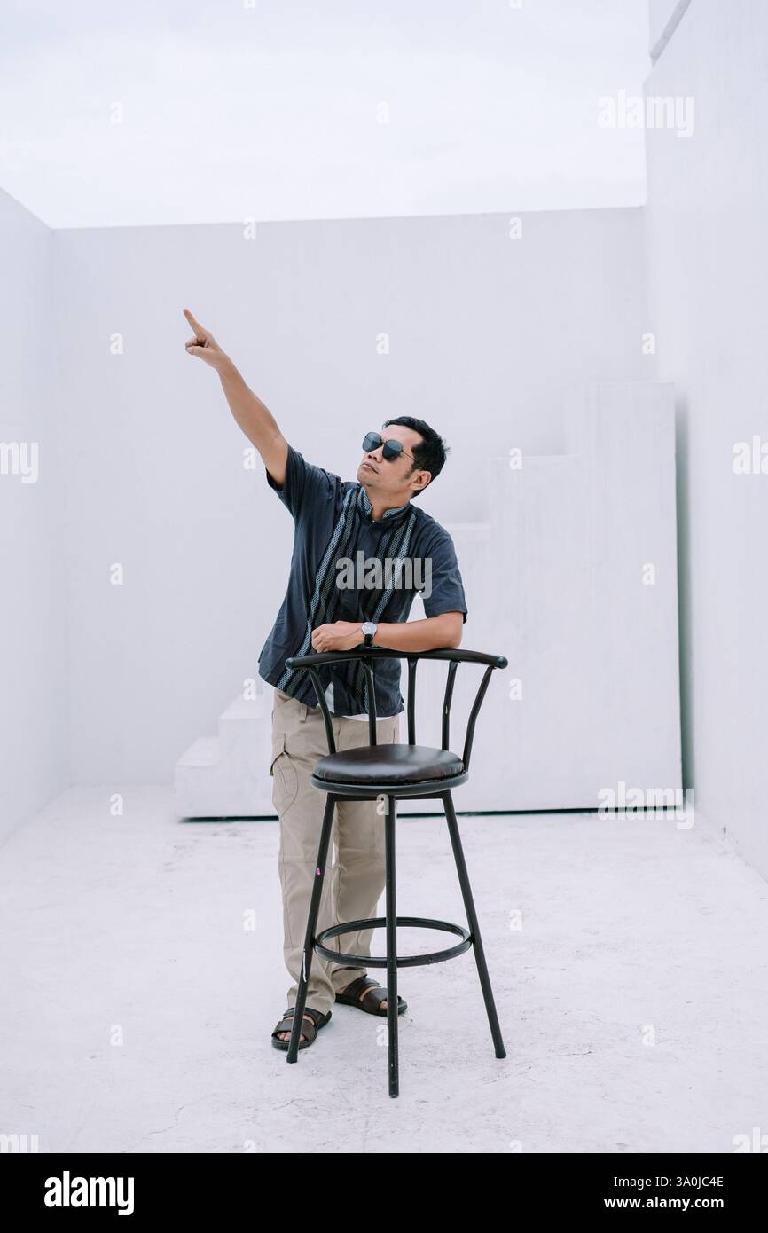 Un elegante uomo asiatico che indossa occhiali da sole punta verso l'alto in uno spazio bianco minimalista. Si appoggia casualmente su una sedia, guardando verso il Foto Stock