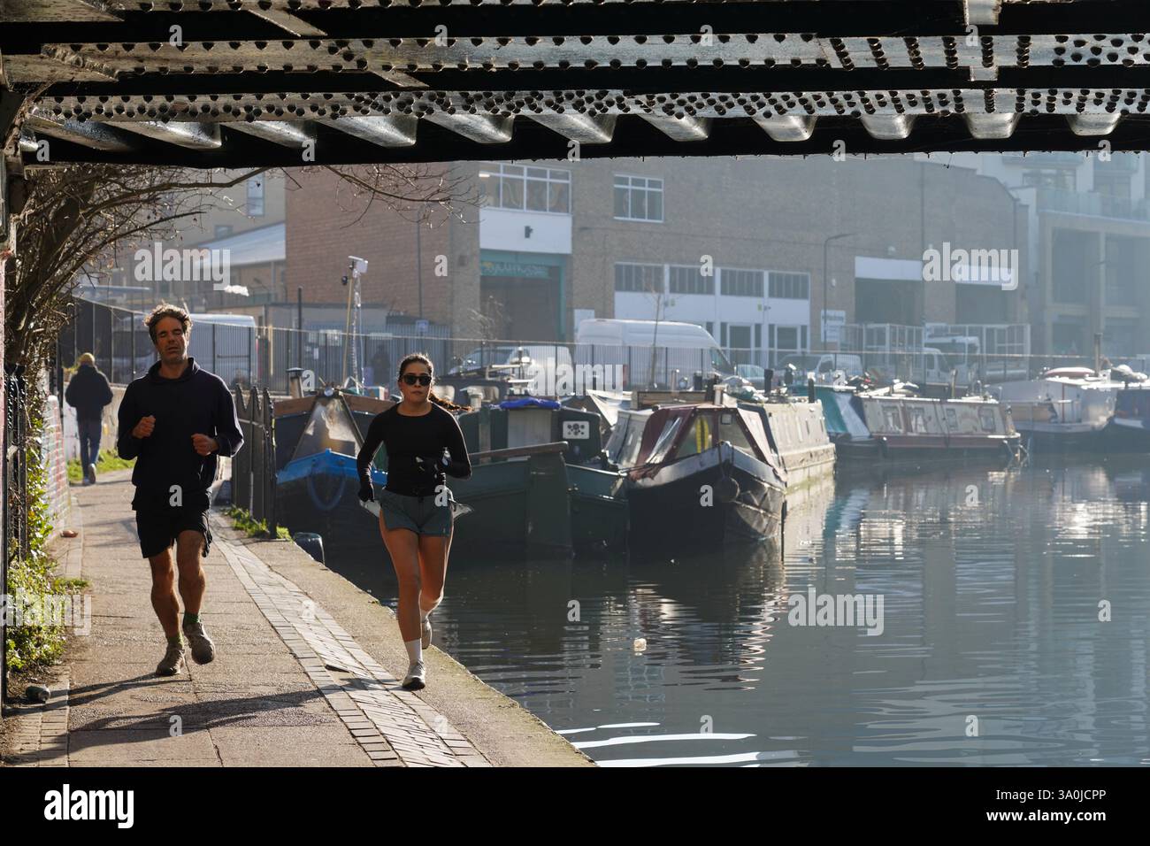 I corridori si allenano lungo il sentiero di traino vicino al Regent's Canal al sole del mattino presto. Le barche della casa costeggiano il canale con il sole che si riflette nell'acqua. Foto Stock