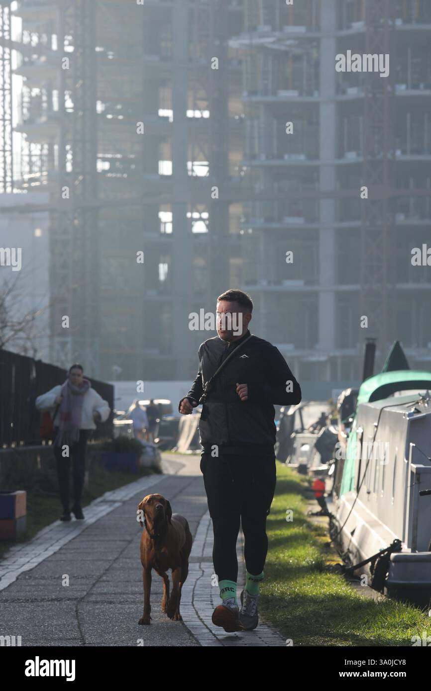 Un corridore si allena lungo il sentiero del Regent's Canal al sole mattutino di primavera. Le barche della casa costeggiano il canale con i lavori di costruzione alle spalle. Foto Stock
