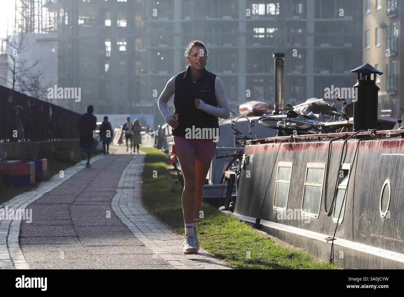 Un corridore si allena lungo il sentiero del Regent's Canal al sole mattutino di primavera. Le barche della casa costeggiano il canale con i lavori di costruzione alle spalle. Foto Stock